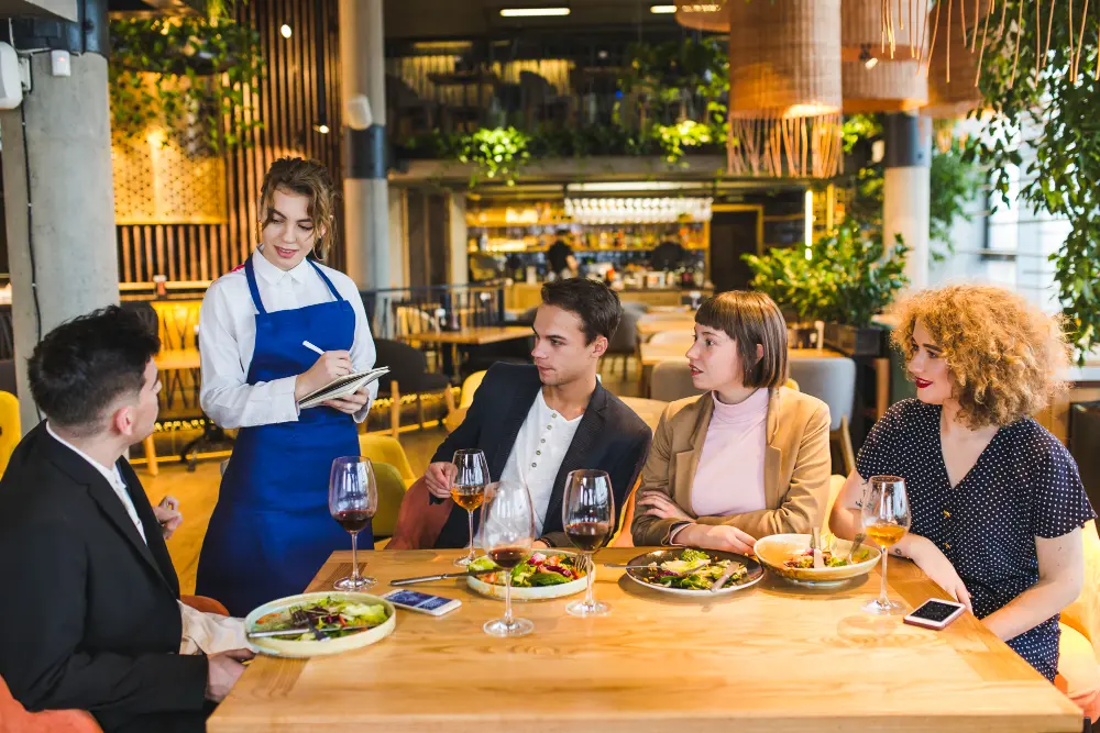 A waitress takes an order from four friends dining at a restaurant table with food, wine, and a lively atmosphere.