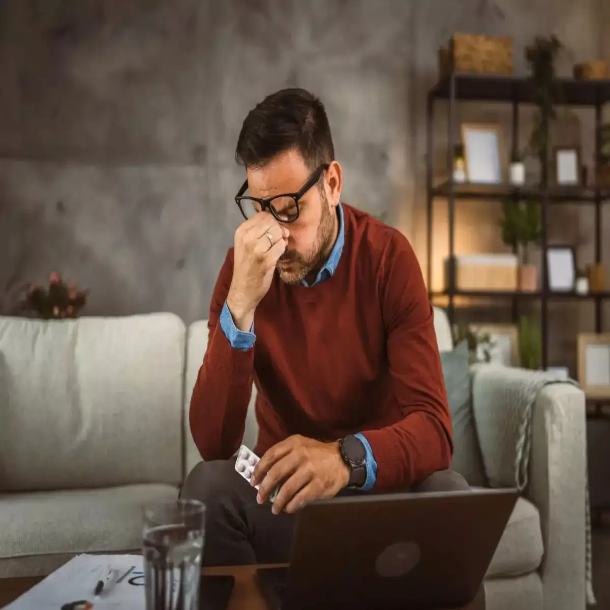 Stressed businessman sitting on a couch holding medicine, facing work challenges while using a laptop at home in Bridgeport.