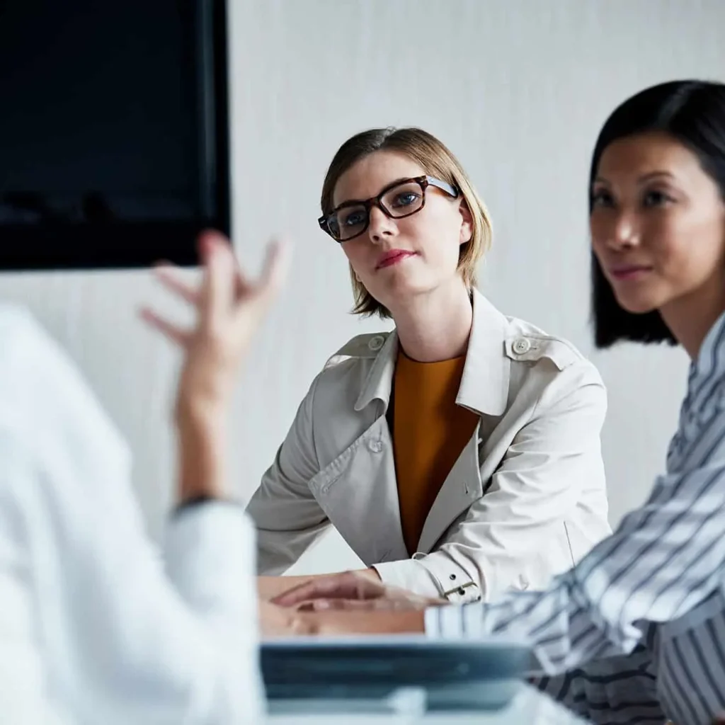 Two women attentively listening to a colleague during a business meeting in a modern office setting.