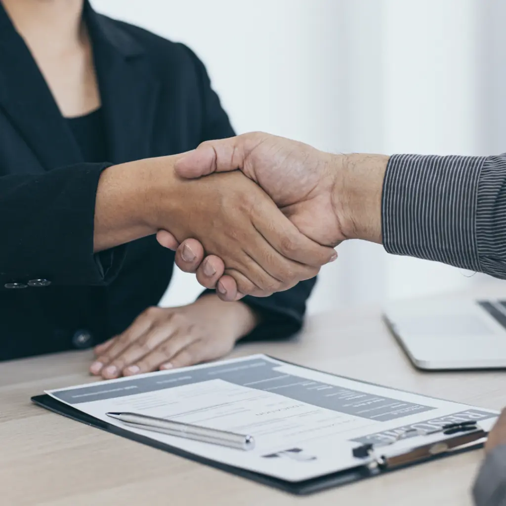 Two people in business attire shake hands across a desk with documents and a pen, symbolizing agreement or partnership.