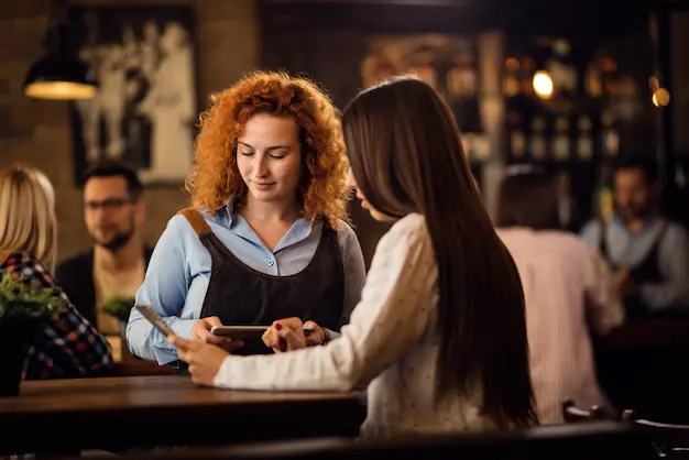 A woman with curly hair holds a payment device while another woman makes a digital payment in a cozy café setting