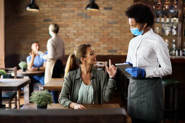 Smiling woman placing an order with a waitress wearing mask and gloves inside a modern cafe with brick wall interior