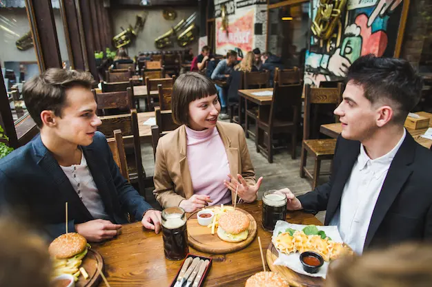 Three friends sitting at a wooden table in a cozy restaurant, enjoying burgers, fries, drinks, and lively conversation