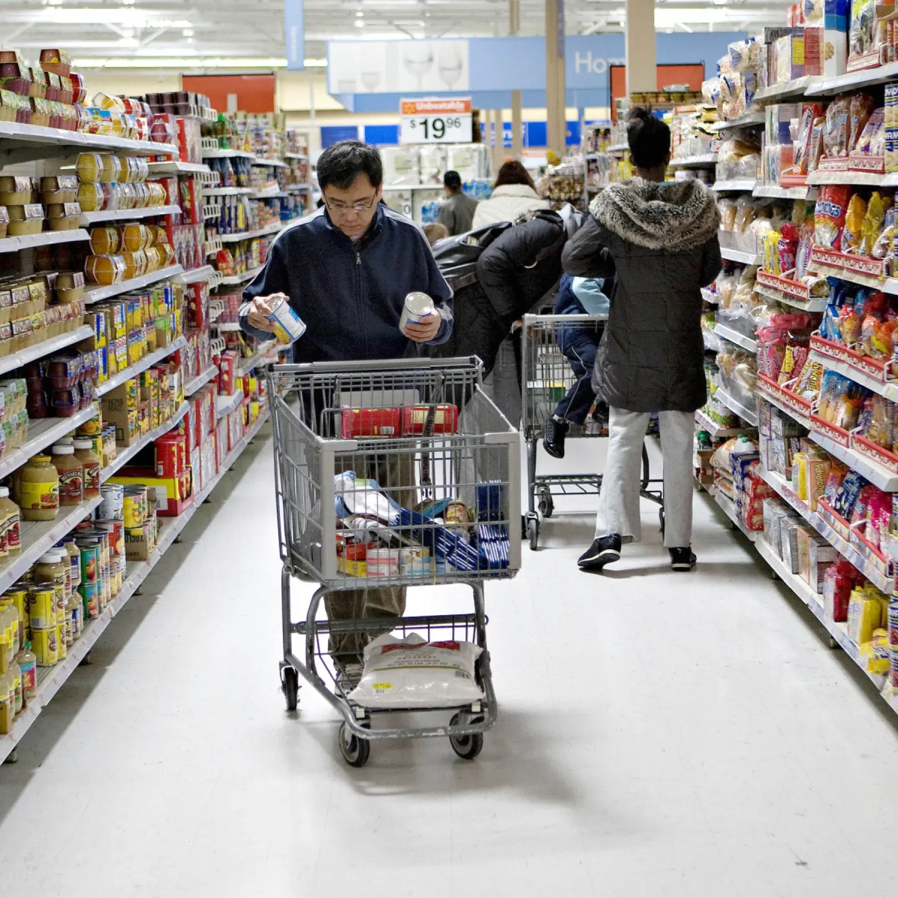 Shoppers browse grocery store aisles, comparing products and filling carts with food and household items.