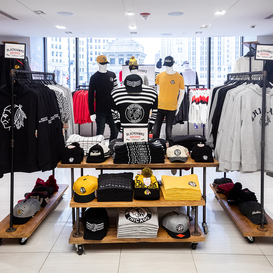 Clothing store display with hoodies, shirts, and hats featuring sports team logos, neatly arranged on racks and tables