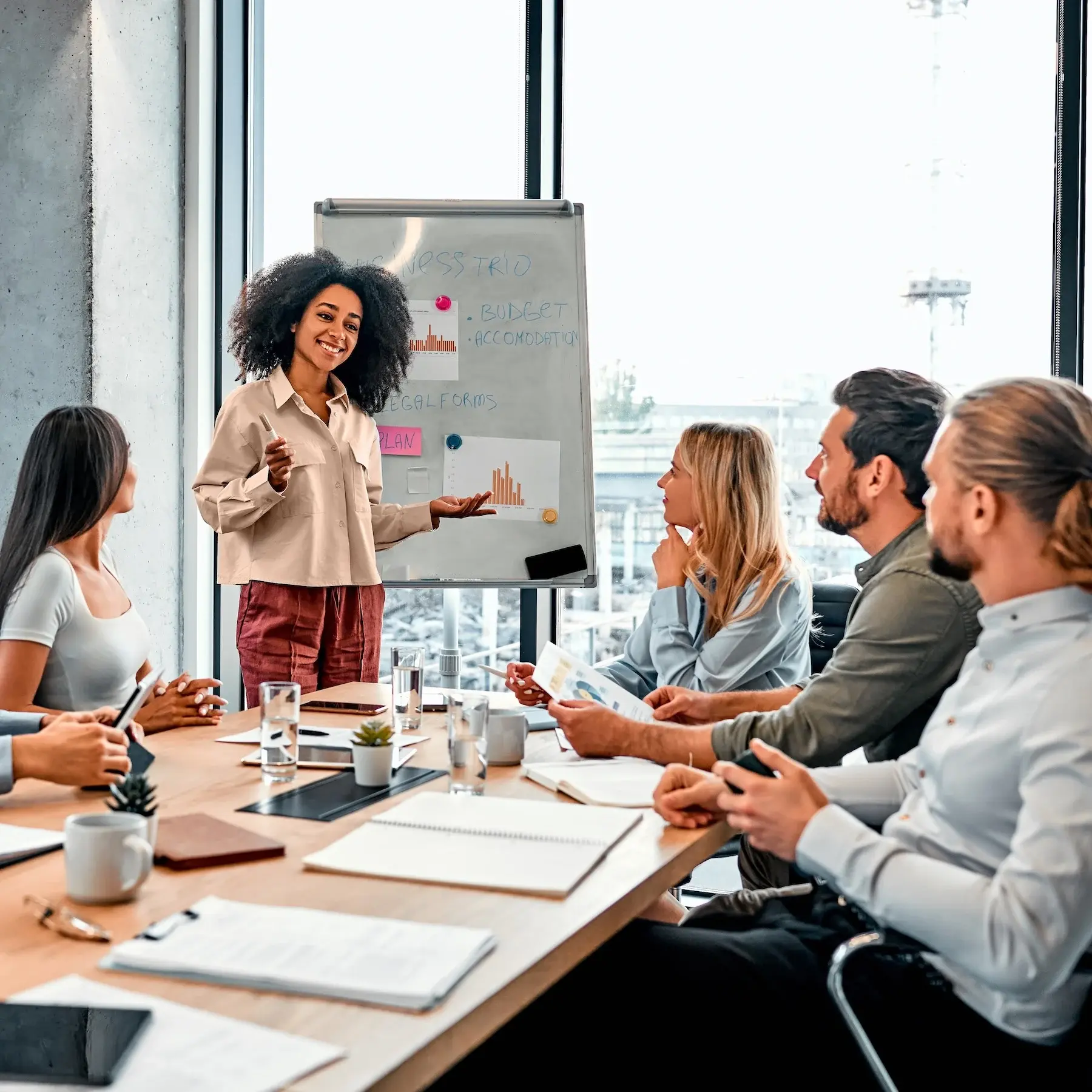 Business team in a meeting with a presenter explaining charts and strategies on a whiteboard in a modern office.