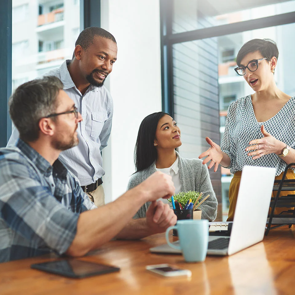 A group of professionals having a discussion around a table with a laptop, tablet, and coffee mugs in a bright modern office