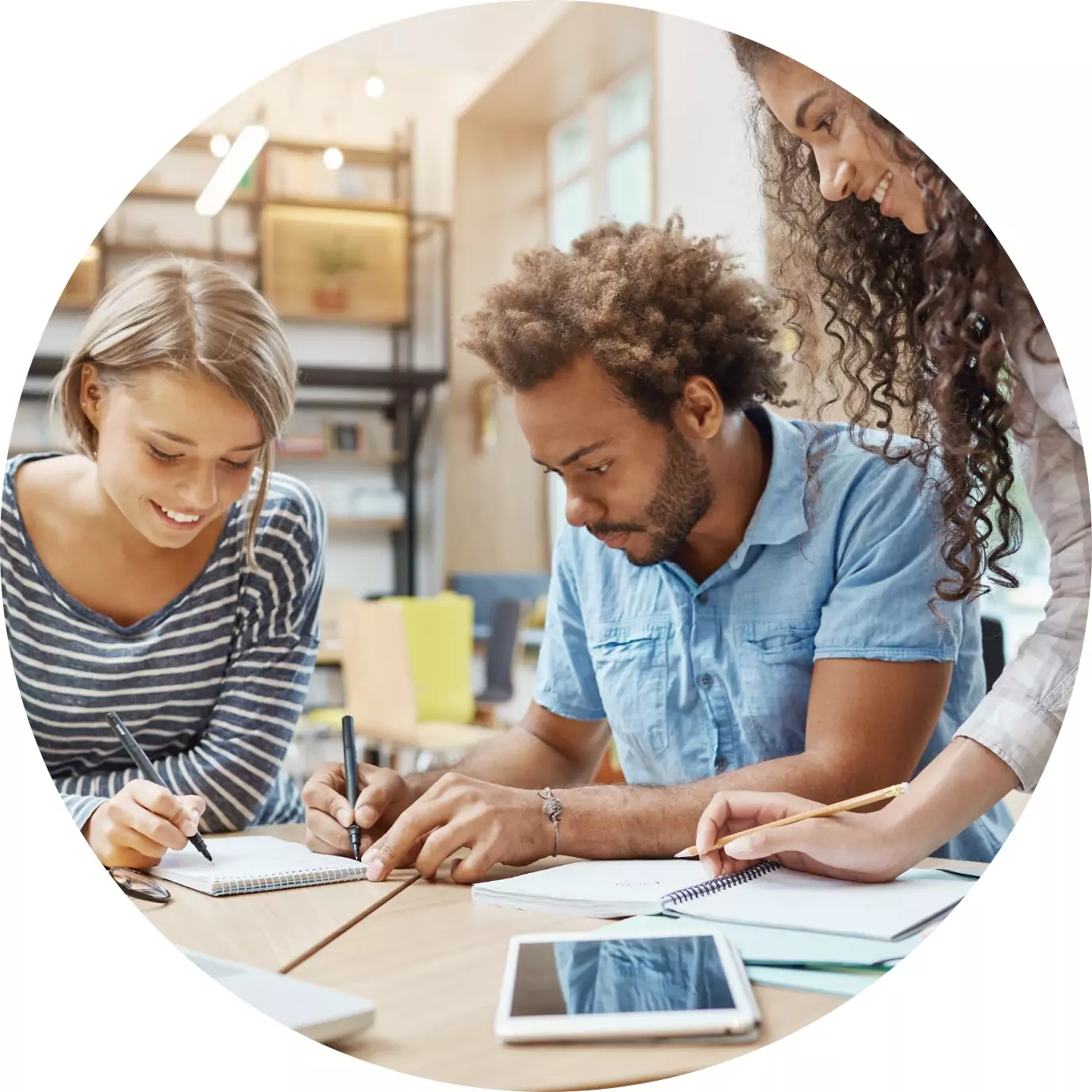 Three people collaborate at a desk, writing in notebooks and smiling in a bright, modern workspace with shelves and a tablet on the table.