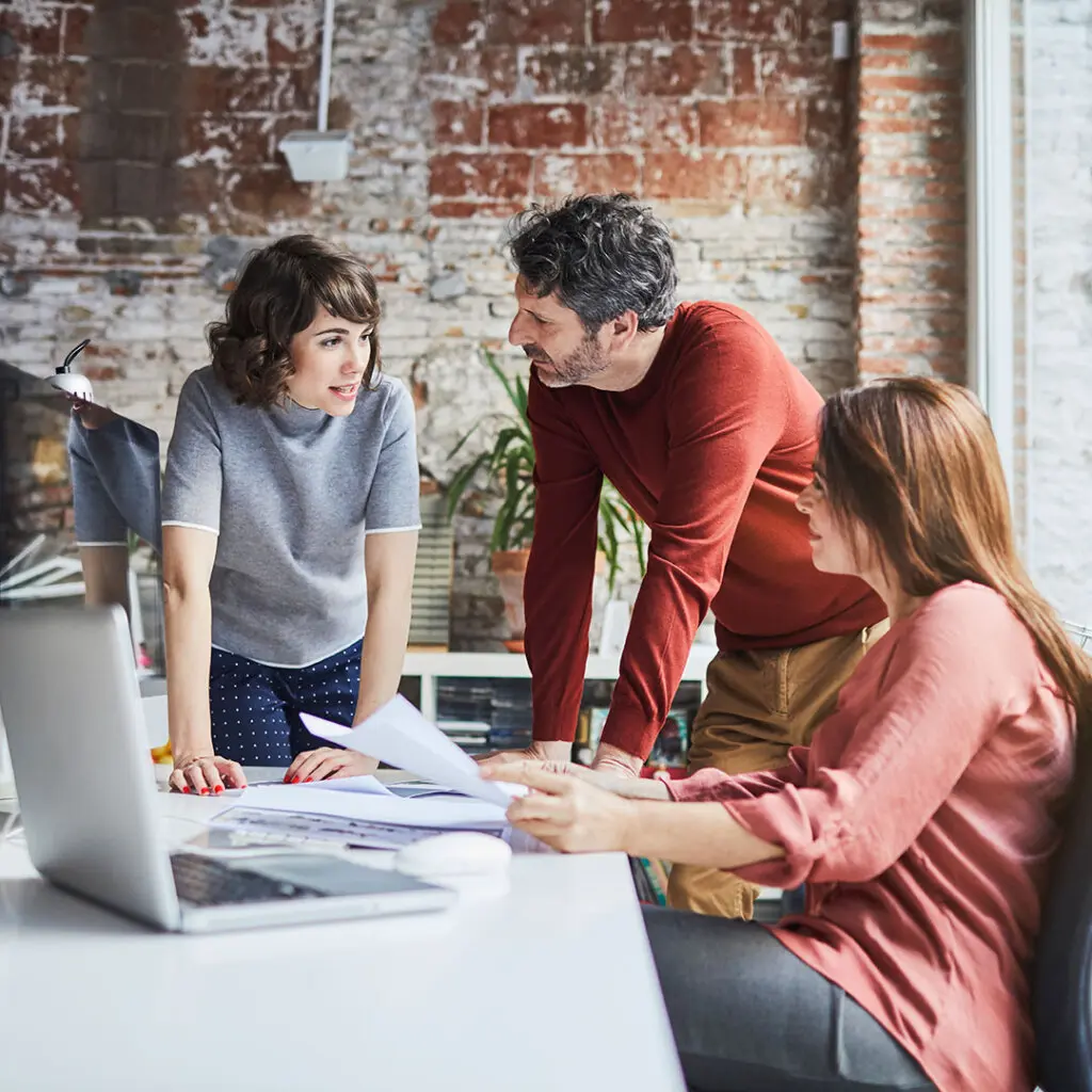 Three professionals discussing documents in a modern office with a laptop, collaborating on business strategies and planning