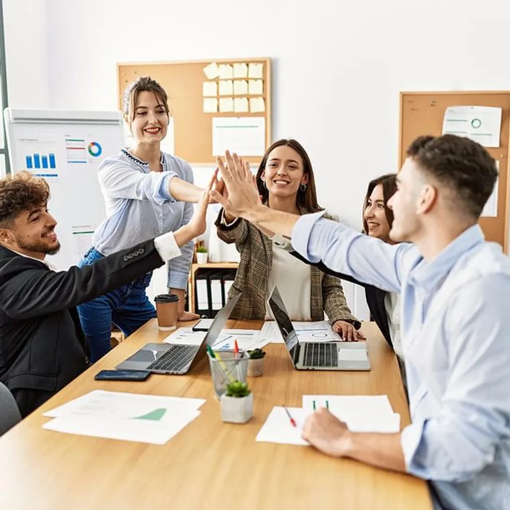 Team of professionals celebrating success with a group high-five in a modern office during a meeting with laptops and reports.