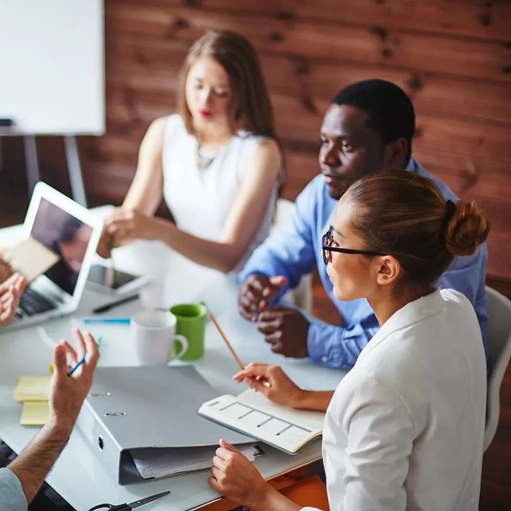 Group of professionals in a meeting room collaborating around a table with laptops, notebooks, and coffee mugs during a business discussion.
