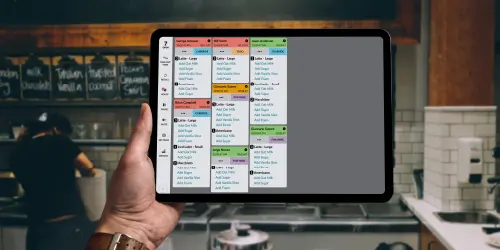 A person holds a tablet showing a digital restaurant order management system in a kitchen with staff working in the background