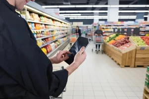Employee using a tablet to manage inventory in a supermarket aisle with fresh produce and shoppers in the background.